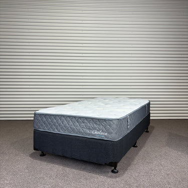 Mattress with 'Chelsea' branding on a gray floor against a corrugated metal wall.