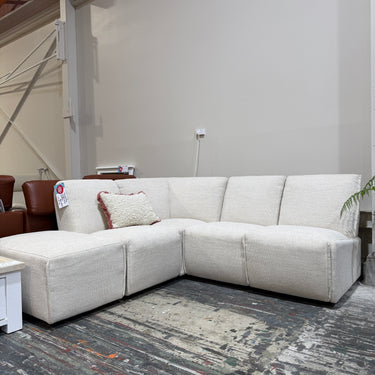 A light beige textured fabric sectional sofa with a left-facing chaise lounge and a small decorative throw pillow, set in a showroom on a distressed gray floor.