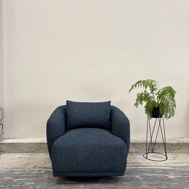 Blue armchair in a room with a plant on a stand against a plain wall.