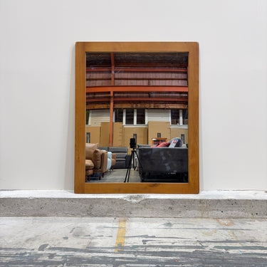 Wooden-framed mirror reflecting a room with furniture on a white wall.