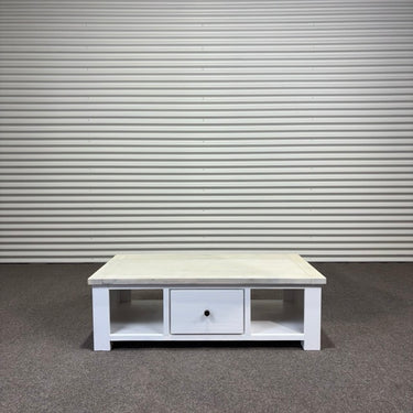 White wooden coffee table with a drawer against a gray corrugated metal wall.