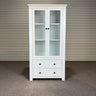 White cabinet with glass doors and drawers against a corrugated metal wall.