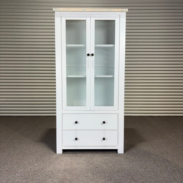 White cabinet with glass doors and drawers against a corrugated metal wall.
