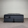 Front view of Backcare Premium mattress with matching base, showing grey quilted top, dark upholstered sides, caster wheels, and Backcare Premium branding on side panel against corrugated backdrop.