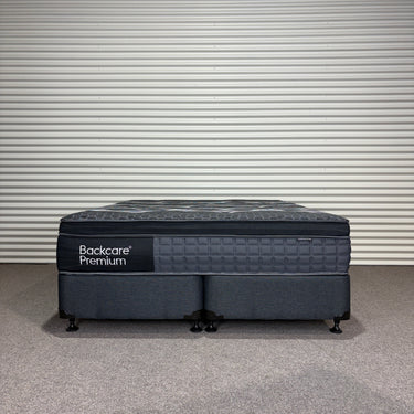 Front view of Backcare Premium mattress with matching base, showing grey quilted top, dark upholstered sides, caster wheels, and Backcare Premium branding on side panel against corrugated backdrop.