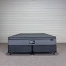 Workhorse King size mattress with matching base, front view showing grey quilted top, dark upholstered base with twin wheel legs and Hotel Mattress Co label against corrugated backdrop