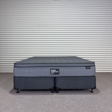 Workhorse King size mattress with matching base, front view showing grey quilted top, dark upholstered base with twin wheel legs and Hotel Mattress Co label against corrugated backdrop