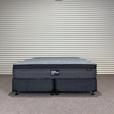 Workhorse LUX mattress king size with matching base, front view showing grey quilted top, dark upholstered base and caster feet against corrugated backdrop