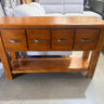 A medium brown wooden console table with four drawers, metal handles, and a lower shelf, photographed in a furniture store setting.
