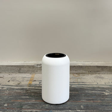 White cylindrical trash can with black lid on a wooden floor against a plain wall.