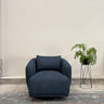 Blue armchair in a room with a plant on a stand against a plain wall.