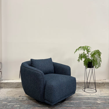 Blue armchair in a room with a plant on a stand against a plain wall.