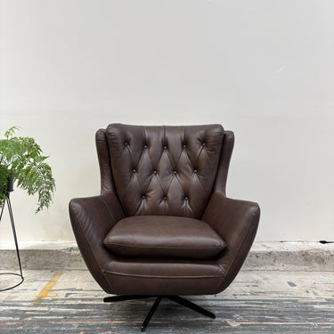 Brown leather armchair with tufted backrest on a wooden floor with a plant in the background.