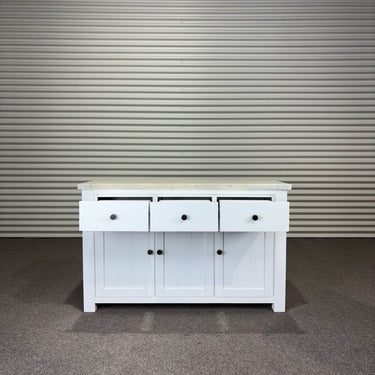 White wooden sideboard with three drawers and two doors against a corrugated metal wall.