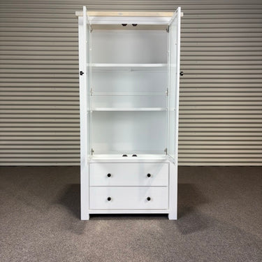 White cabinet with glass doors and drawers against a corrugated metal wall.