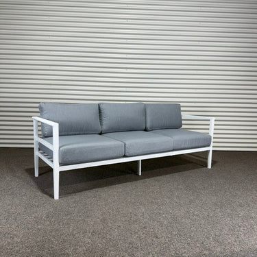 Modern outdoor sofa with gray cushions on a white metal frame against a corrugated metal wall.