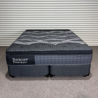 Front view of Backcare Premium mattress with matching base showing grey quilted top, dark upholstered sides, standard legs and branded side panel against corrugated backdrop.