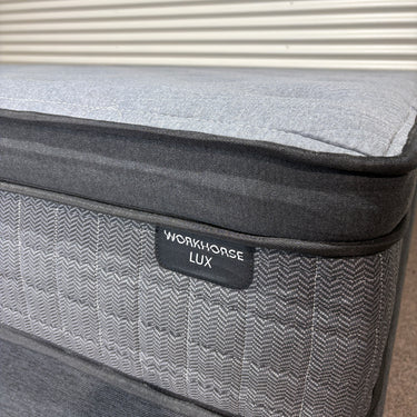 Close-up of Workhorse LUX mattress corner showing grey textured fabric with herringbone pattern, black piping, and Workhorse LUX label, highlighting stitching details.