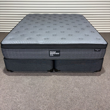 Front top view of Workhorse LUX mattress with matching base showing grey quilted top, dark upholstered base and Hotel Mattress Co branding against corrugated backdrop