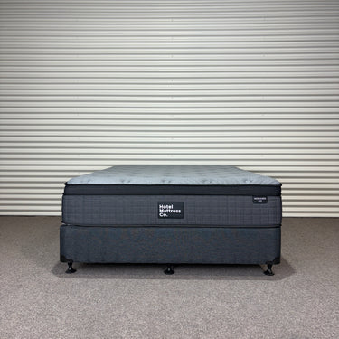 Workhorse LUX mattress with matching base, front view showing quilted top, dark upholstered fabric and brand label against corrugated backdrop